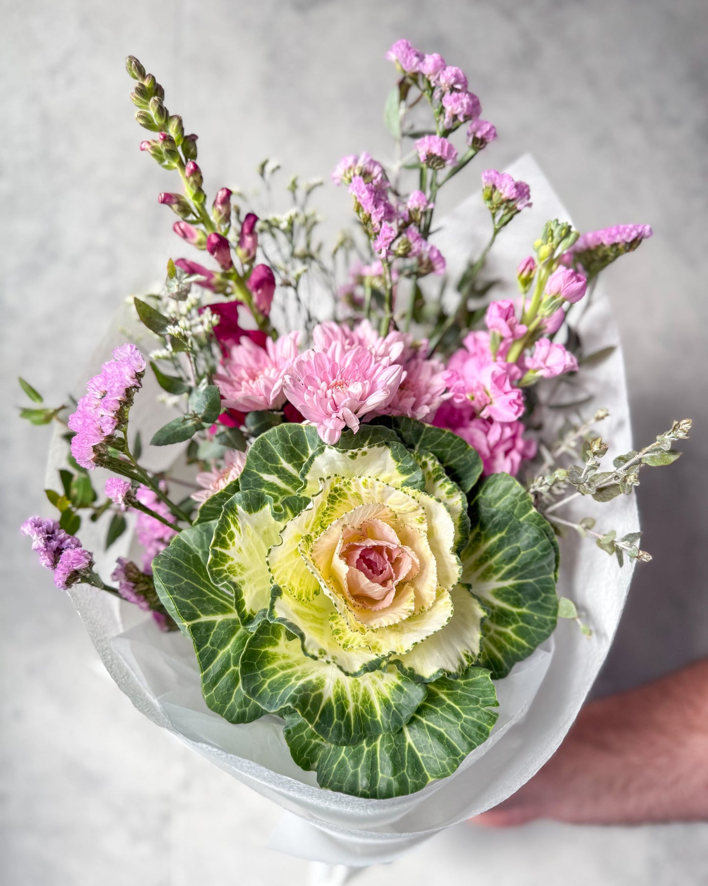 A small seasonal posy featuring a big beautiful pink and white kale, mauve flowers and a plum snapdragon wrapped in white paper.